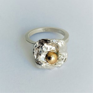 Op de afbeelding: Een zilveren ring met een getextureerde, organisch gevormde zetting. Een kleine, ronde, gouden kraal bevindt zich in het midden van de zetting.