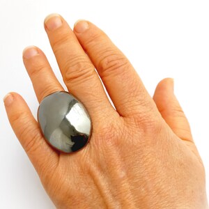 Puede incluir: Un anillo plateado grande, de forma ovalada. La superficie del anillo es lisa y reflectante. El anillo se lleva en un dedo, destacando su tamaño y diseño. El anillo es una pieza llamativa, probablemente de metal. Mide aproximadamente 3 cm de largo.