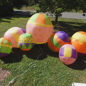 May include: A collection of six inflatable egg-shaped decorations in various sizes, featuring vibrant patterns of orange, pink, green, yellow, and purple. The eggs display geometric designs including stripes, polka dots, and triangles, with a white power cord.