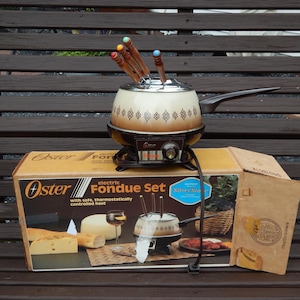May include: An Oster electric fondue set with a cream and brown pot featuring a geometric pattern, a black base with a temperature control dial, and four fondue forks with wooden handles and colored tips, sitting on its original cardboard box.