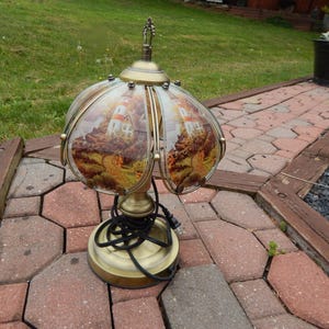 May include: A vintage brass table lamp with a glass shade featuring a scenic lighthouse design. The lamp has a round base, a curved stem, and a decorative finial. The shade depicts a lighthouse scene with warm, earthy tones.