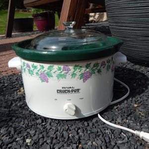 May include: A white Rival Crock-Pot slow cooker with a glass lid and green trim. The slow cooker has a floral design with purple grapes and green leaves. It has a white power cord and a three-setting dial.