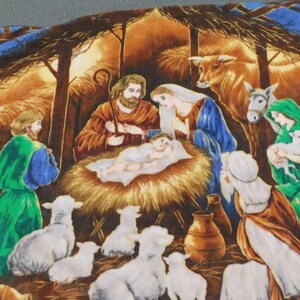 May include: A rectangular fabric panel featuring a colorful nativity scene illustration. The print depicts a central manger with a baby, surrounded by figures in robes, a cow, a donkey, and several white lambs under a thatched roof structure.