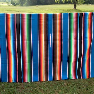 May include: A rectangular serape blanket featuring a vibrant, multi-colored striped pattern. The design includes bold vertical bands of bright blue, deep brown, black, green, red, orange, and yellow, finished with white fringe on both ends.