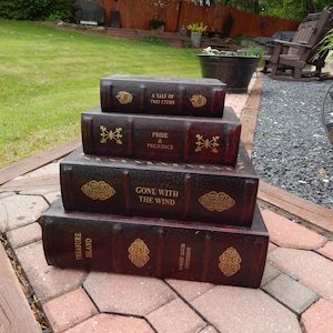 May include: A stack of four decorative book boxes in varying sizes. The boxes are dark brown with gold-colored lettering and embellishments. The titles include "Treasure Island," "A Tale of Two Cities," "Pride & Prejudice," and "Gone With The Wind."