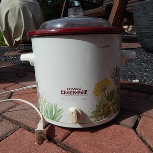 May include: White Rival Crock-Pot slow cooker with a clear glass lid and a maroon rim. The slow cooker has a floral design with green and yellow accents. It has a white power cord and a control dial with settings for Off, Low, and High.