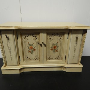 May include: Cream-colored jewelry box with floral accents. The box has two doors with gold-toned handles and decorative floral designs. The sides feature vertical detailing and gold-toned hardware. The top is flat and slightly overhanging.