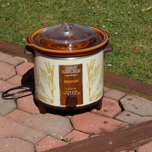 May include: A vintage Crock Watcher slow cooker with a cream-colored body, brown trim, and a clear glass lid. The front features a wood-grain panel with a control knob and wheat stalk illustrations. It includes a black power cord and side handles.