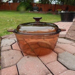 May include: A transparent amber-colored casserole dish with a matching lid. The dish has two side handles and a knob on the lid. The dish is sitting on a brick patio.