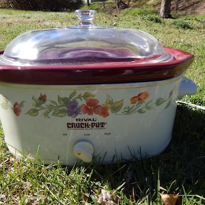 May include: A Rival Crock-Pot stoneware slow cooker with a clear glass lid and a burgundy rim. The cream-colored pot features a floral design and a dial with settings for off, low, and high.