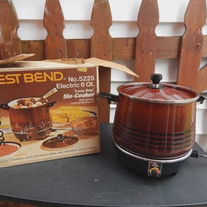 May include: A vintage West Bend Slo-Cooker, model No. 5225, with a 6-quart capacity. The electric slow cooker has a brown exterior with black stripes and a black base. The original box is also present.