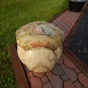 May include: A round upholstered ottoman featuring a top cushion with a woven tapestry scene of figures in a landscape. The base is covered in a cream-colored fabric with a subtle tone-on-tone damask pattern and vertical pleats around the sides.
