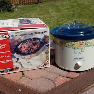 May include: A white Rival Crock-Pot with a blue lid and floral design, next to its original box. The box reads "6 Quart" and "Olla de cocimiento lento CRUCK-POT". The slow cooker has a white dial and handles.