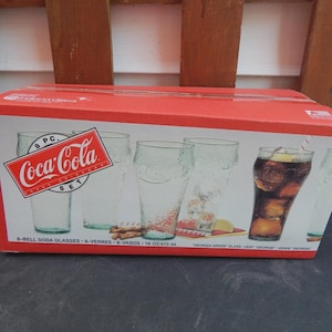 May include: A red cardboard box containing an 8-piece Coca-Cola soda glass set. The box features images of clear, textured bell-shaped glasses, one filled with a dark beverage and ice. Text on the box reads 8 PC. Coca-Cola SET and 16 oz/473 ml.