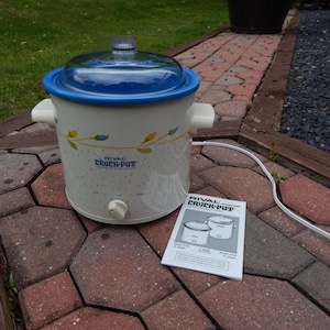May include: A white Rival Crock-Pot slow cooker with a blue rim and lid, and a floral design. The appliance has a white power cord and a control knob. A manual is next to the slow cooker.