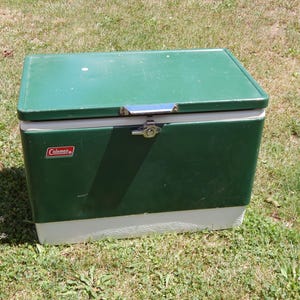 May include: A rectangular Coleman cooler featuring a dark green metal exterior with a white base. The lid includes a silver-toned metal latch mechanism centered on the front, and the Coleman logo is printed in red and white on the upper left side.