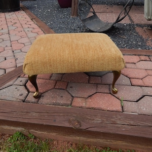 May include: A rectangular footstool featuring a textured, mustard-yellow fabric upholstery. The piece is supported by four curved, metallic gold-toned legs that terminate in rounded feet, providing a stable base for the cushioned top surface.