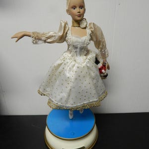 May include: A porcelain ballerina doll in a white dress with gold polka dots and trim. The doll has blonde hair, a gold crown, and a gold choker. It stands on a blue and cream-colored base. The doll is holding a small object.