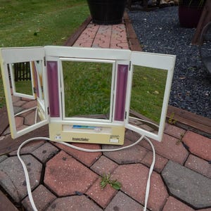 May include: A vintage Image-Maker vanity mirror with three hinged panels. The mirror has a cream-colored base with a white frame and purple accent lights. A white power cord extends from the base. The mirror is open, reflecting the surrounding environment.