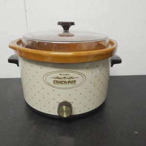 May include: A Rival Crock-Pot slow cooker with a brown and cream color scheme. The appliance has a glass lid, brown handles, and a control knob. The Crock-Pot is decorated with a repeating pattern of small, dark shapes and the words "RIVAL CROCK-POT".