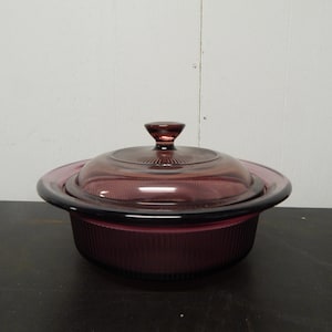 May include: A vintage, round, purple glass casserole dish with a matching lid. The dish has a ribbed design on the sides and a clear glass lid with a knob handle. The dish is sitting on a dark surface.