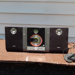 May include: A black and silver Philips stereo system featuring a central CD player unit with a visible disc, flanked by two integrated speakers. The unit includes a small digital display, control buttons, and a separate black and grey remote control.