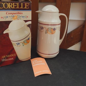 May include: A Corelle Abundance thermal server with a matching lid and handle, alongside its original box. The server is cream-colored with a decorative band featuring fruit and floral designs. A small orange card is also present.