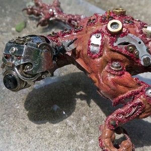 Könnte beinhalten: Nahaufnahme einer roten, fleischigen und metallischen Skulptur einer Kreatur. Die Kreatur hat einen metallischen Kopf mit Zahnrädern und einer Kamera-Linse. Der Körper ist mit roter Farbe bedeckt und hat viele daran befestigte Metallteile.