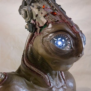 May include: A close-up of a brown, sculpted bust of an alien with a single, glowing blue eye. The alien's head is topped with a complex, steampunk-style mechanical device.