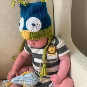 May include: A baby wearing a blue, green, and yellow knitted owl hat with a lion patch on their grey and white striped onesie. The baby is holding a blue knitted fish toy.