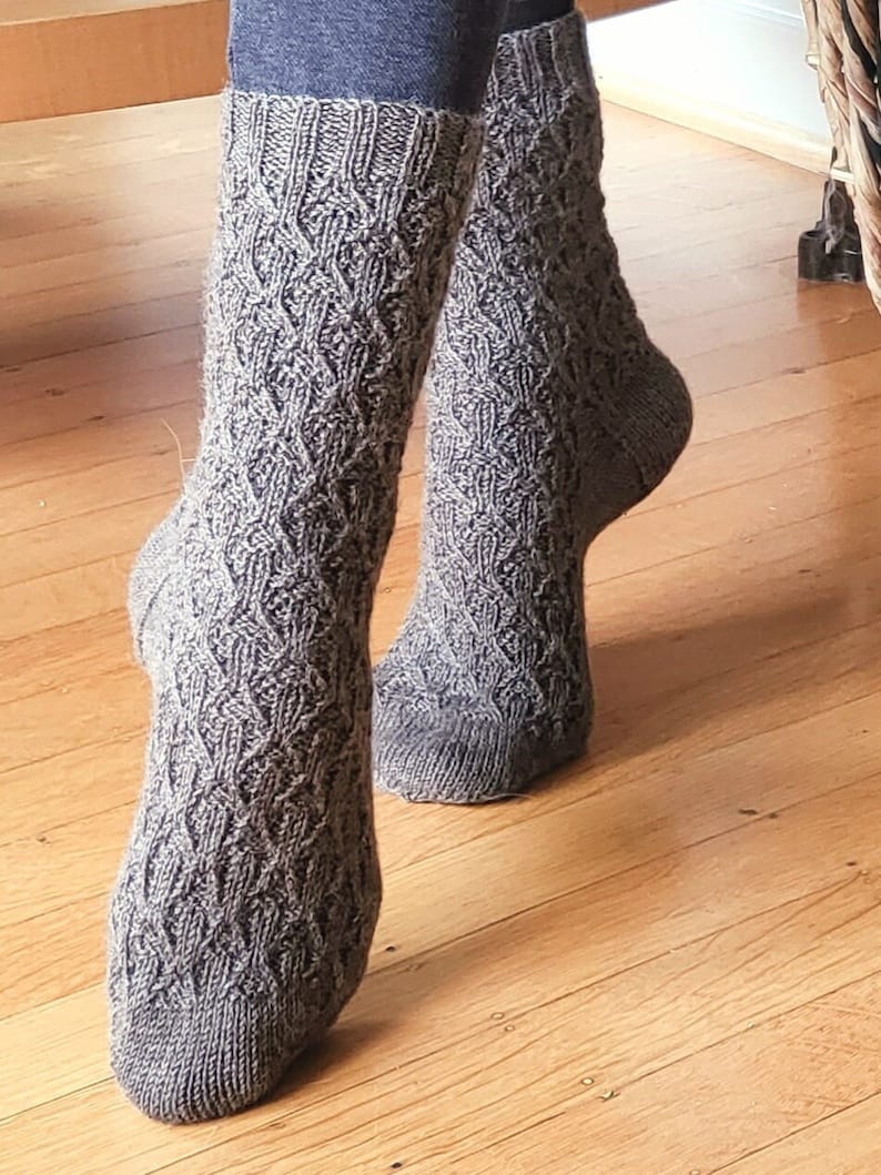 May include: A pair of gray knitted socks with a cable knit pattern. The socks are worn on a person's feet.