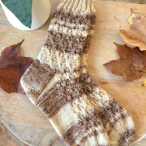 May include: A pair of brown and cream knitted socks with a cable knit pattern. The socks are lying on a wooden surface.