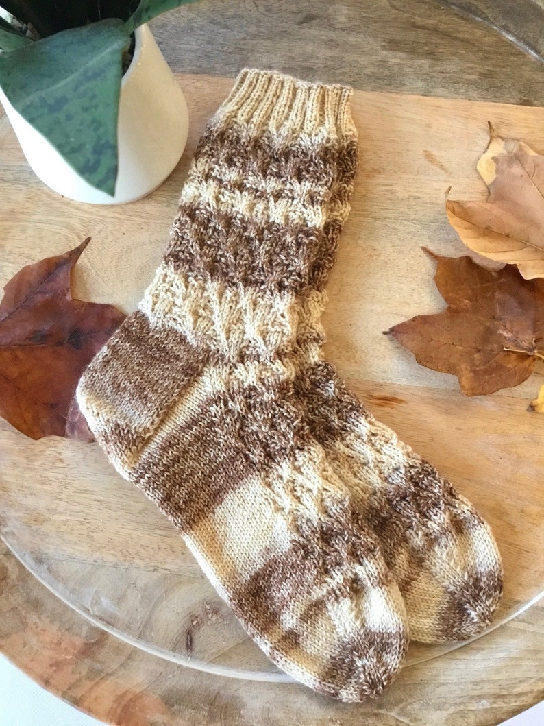 May include: A pair of brown and cream knitted socks with a cable knit pattern. The socks are lying on a wooden surface.
