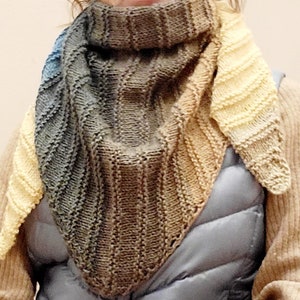 K&ouml;nnte beinhalten: Ein dreieckiger gestrickter Schal mit einem Farbverlauf von Blau, Braun und Gelb. Der Schal ist gefaltet und wird von einer Person um den Hals getragen.