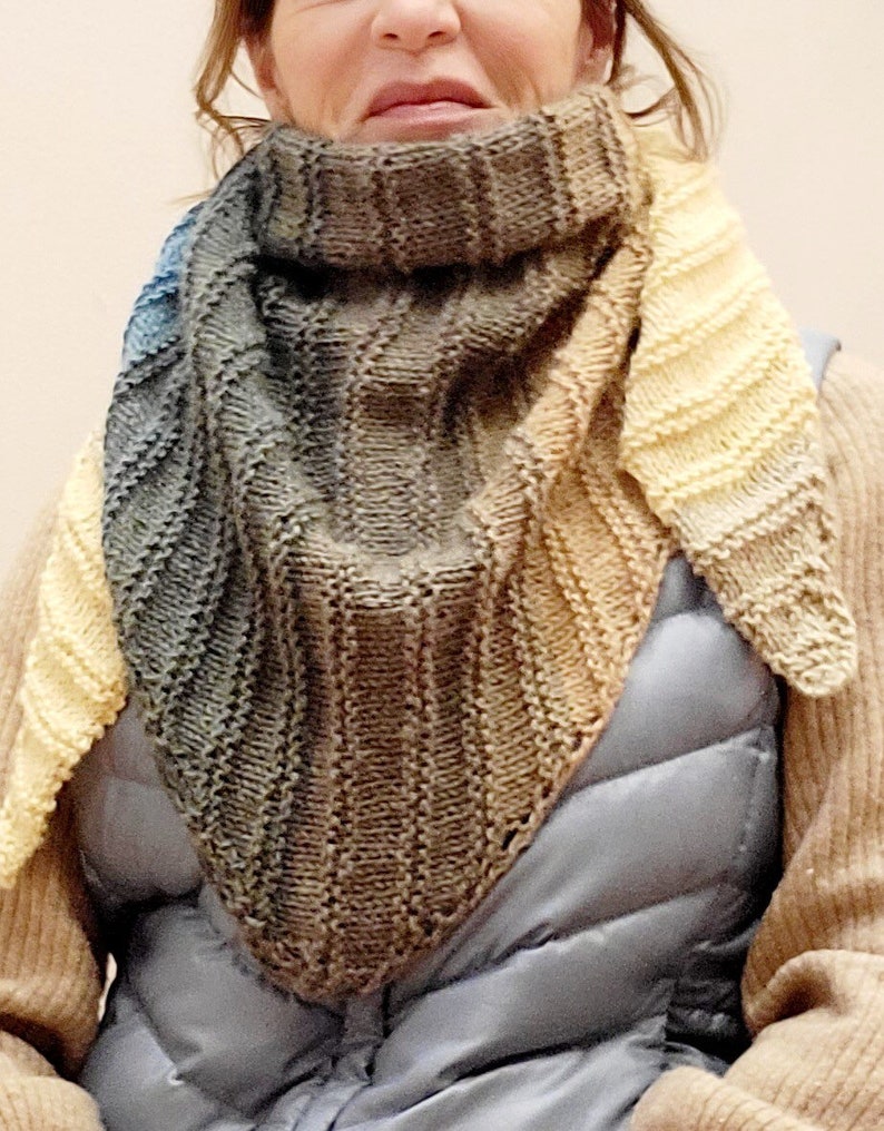 K&ouml;nnte beinhalten: Ein dreieckiger gestrickter Schal mit einem Farbverlauf von Blau, Braun und Gelb. Der Schal ist gefaltet und wird von einer Person um den Hals getragen.