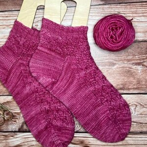 May include: Two pink knitted socks with a cable pattern, displayed on wooden sock blockers. A ball of pink yarn is also shown.