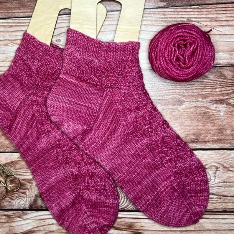 May include: Two pink knitted socks with a cable pattern, displayed on wooden sock blockers. A ball of pink yarn is also shown.