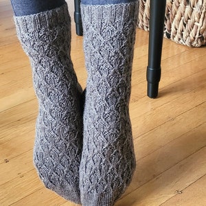 May include: A pair of gray knitted socks with a cable knit pattern. The socks are worn on a person's feet and are folded down at the top.