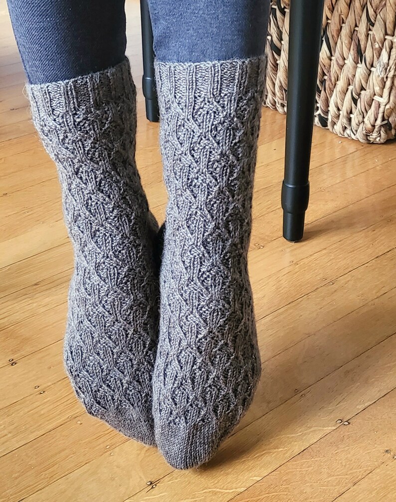 May include: A pair of gray knitted socks with a cable knit pattern. The socks are worn on a person's feet and are folded down at the top.