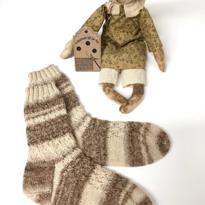 May include: A pair of brown and cream striped knitted socks. The socks are handmade and have a rustic, homespun look. The socks are displayed with a handmade doll wearing a brown and yellow floral dress.