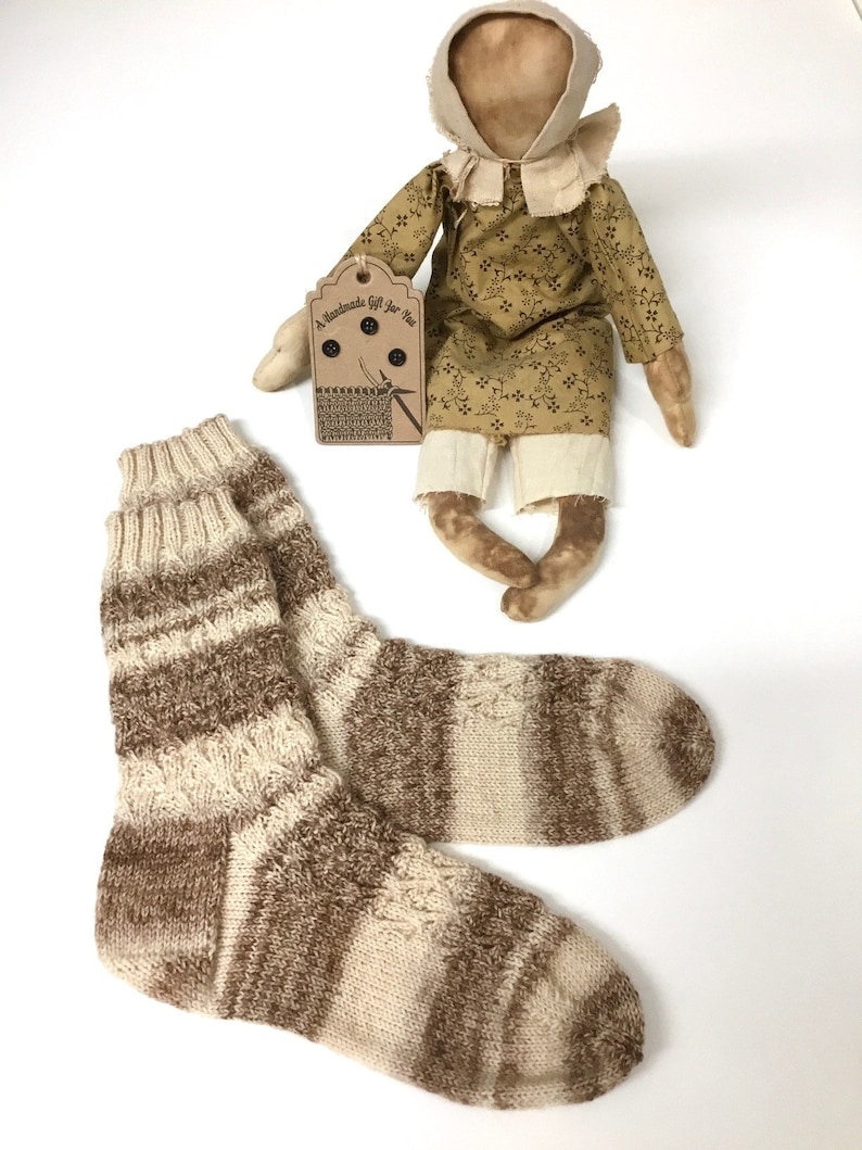 May include: A pair of brown and cream striped knitted socks. The socks are handmade and have a rustic, homespun look. The socks are displayed with a handmade doll wearing a brown and yellow floral dress.