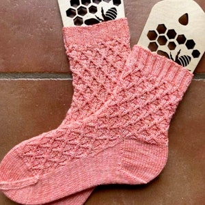 May include: A pair of pink knitted socks with a honeycomb pattern. The socks are displayed on wooden tags with a bee and honeycomb design.