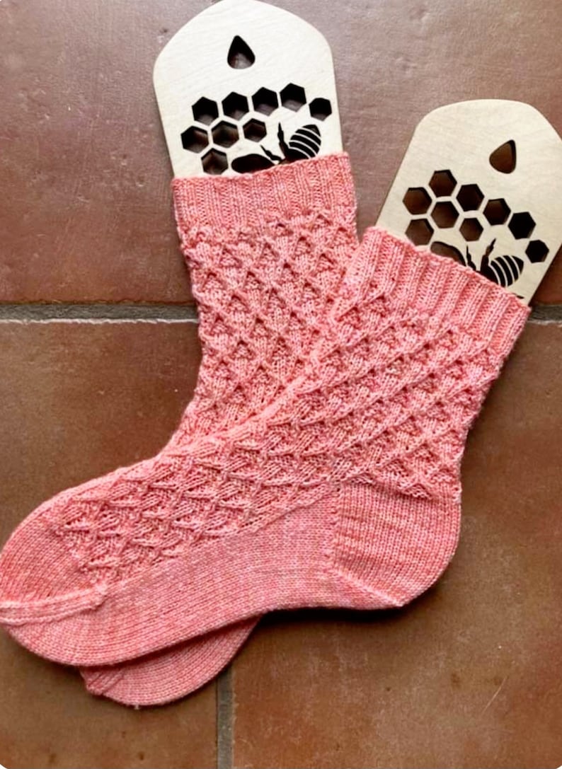 May include: A pair of pink knitted socks with a honeycomb pattern. The socks are displayed on wooden tags with a bee and honeycomb design.