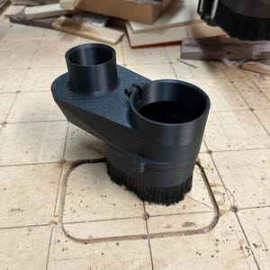Könnte beinhalten: Ein schwarzer, 3D-gedruckter Staubschuh-Aufsatz für eine CNC-Maschine mit zwei zylindrischen Anschlüssen an der Oberseite und einem kreisförmigen Bürstenkranz an der Basis. Das Bauteil hat eine texturierte Oberfläche zur Schmutzaufnahme.