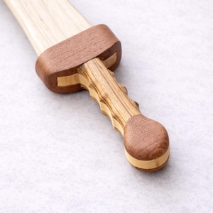 May include: A wooden sword with a light-colored blade and a dark brown handle. The handle has a detailed, carved design and a rounded pommel. The guard is also dark brown, with a lighter wood accent.