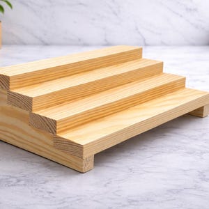 Pode incluir: Uma prateleira de exibição em madeira natural com três níveis. Cada nível é ligeiramente mais alto que o anterior e é suportado por uma base. O grão da madeira é visível e a prateleira não está acabada.