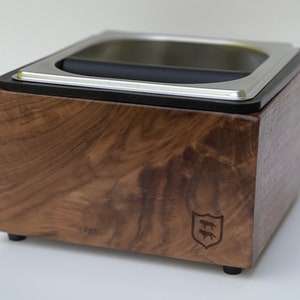 Op de afbeelding: Een houten espresso knock box met een roestvrijstalen bovenkant en een zwarte rubberen bar. De doos is gemaakt van donker hout met een natuurlijk nerfpatroon en een klein embleem aan de zijkant.