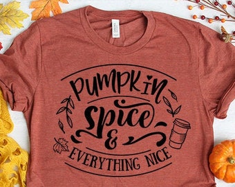 May include: A rust-colored t-shirt with the text "Pumpkin Spice & Everything Nice" in black lettering. The shirt is surrounded by fall-themed decor, including a yellow scarf, jeans, and fall leaves.