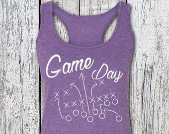 May include: A purple racerback tank top with the words "Game Day" in white script. Below the text is a white graphic of a football play. The tank top has a scoop neck and a racerback design.