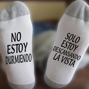 May include: A pair of white socks with black text. The left sock says "No estoy durmiendo" and the right sock says "Solo estoy descansando la vista".